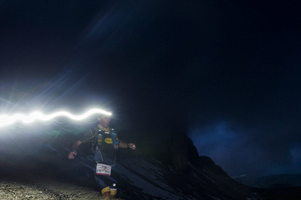 Corridore con lampada frontale accesa di notte in montagna, la scia di luce illumina il sentiero