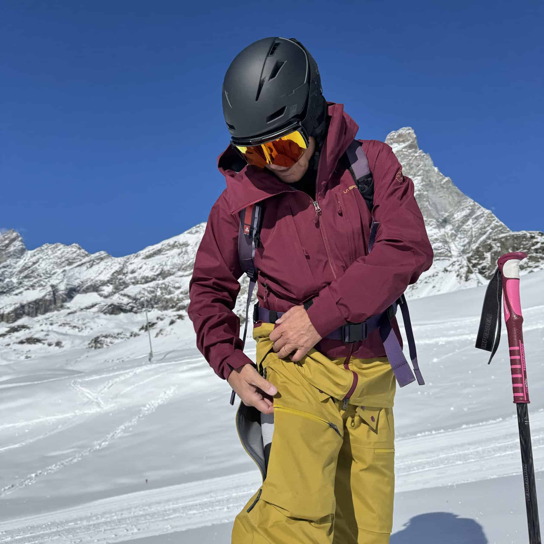 Casco da sci nero con visiera gialla, uomo in abbigliamento da montagna sulla neve, montagne e cielo blu sullo sfondo.