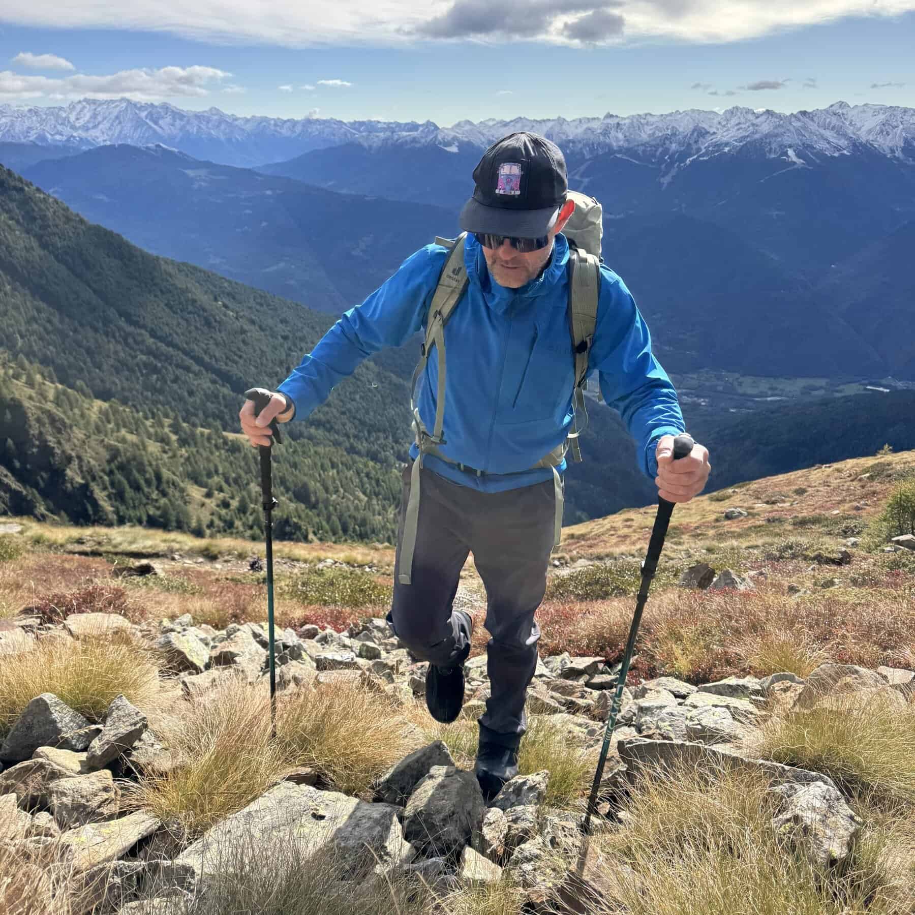 Escursionista con softshell blu, zaino e bastoncini in salita su sentiero alpino roccioso, panorama di montagne innevate — attrezzatura outdoor estate 2026