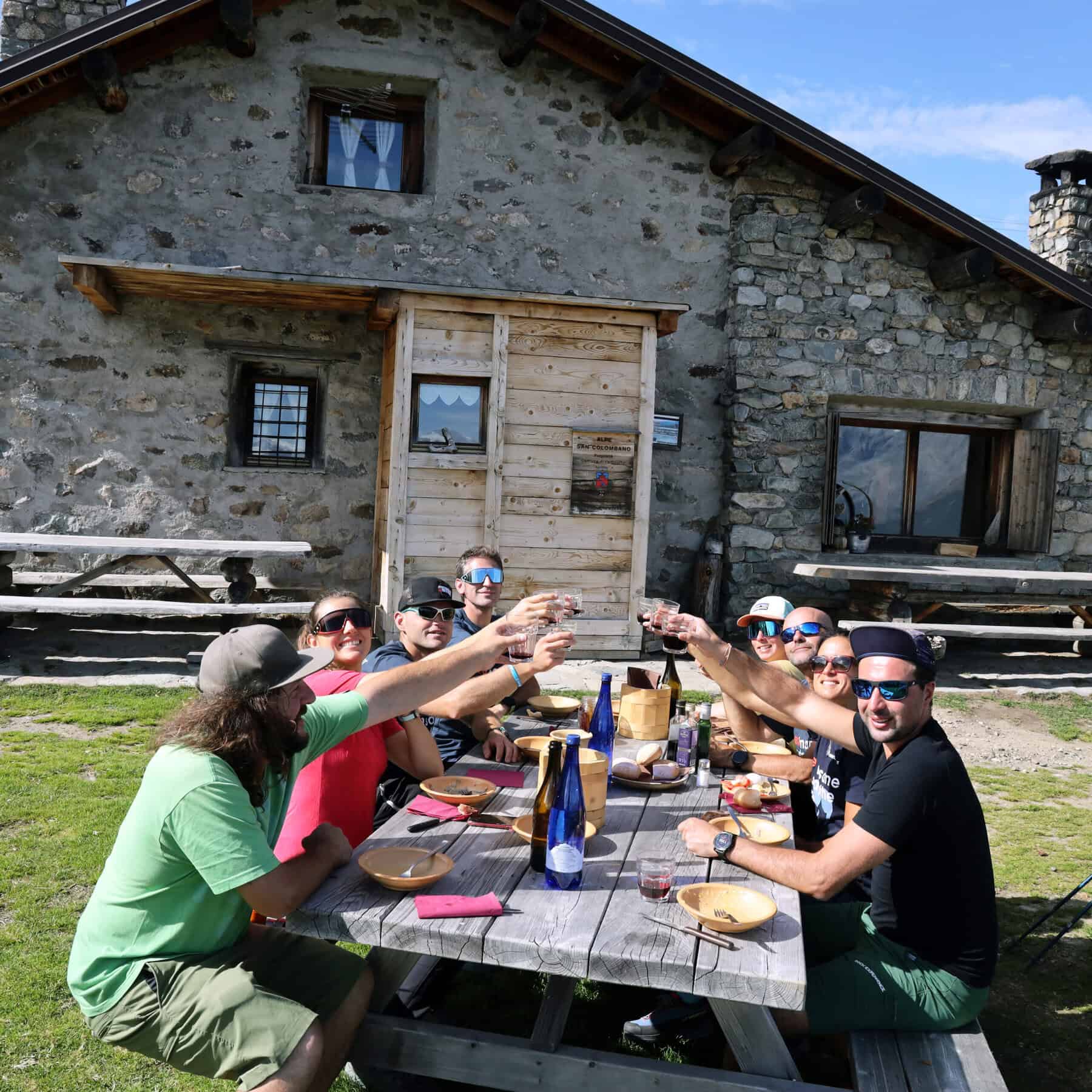 Gruppo di tester brinda a tavola davanti a rifugio alpino in pietra in piena estate — community Outdoortest, checklist outdoor estate 2026
