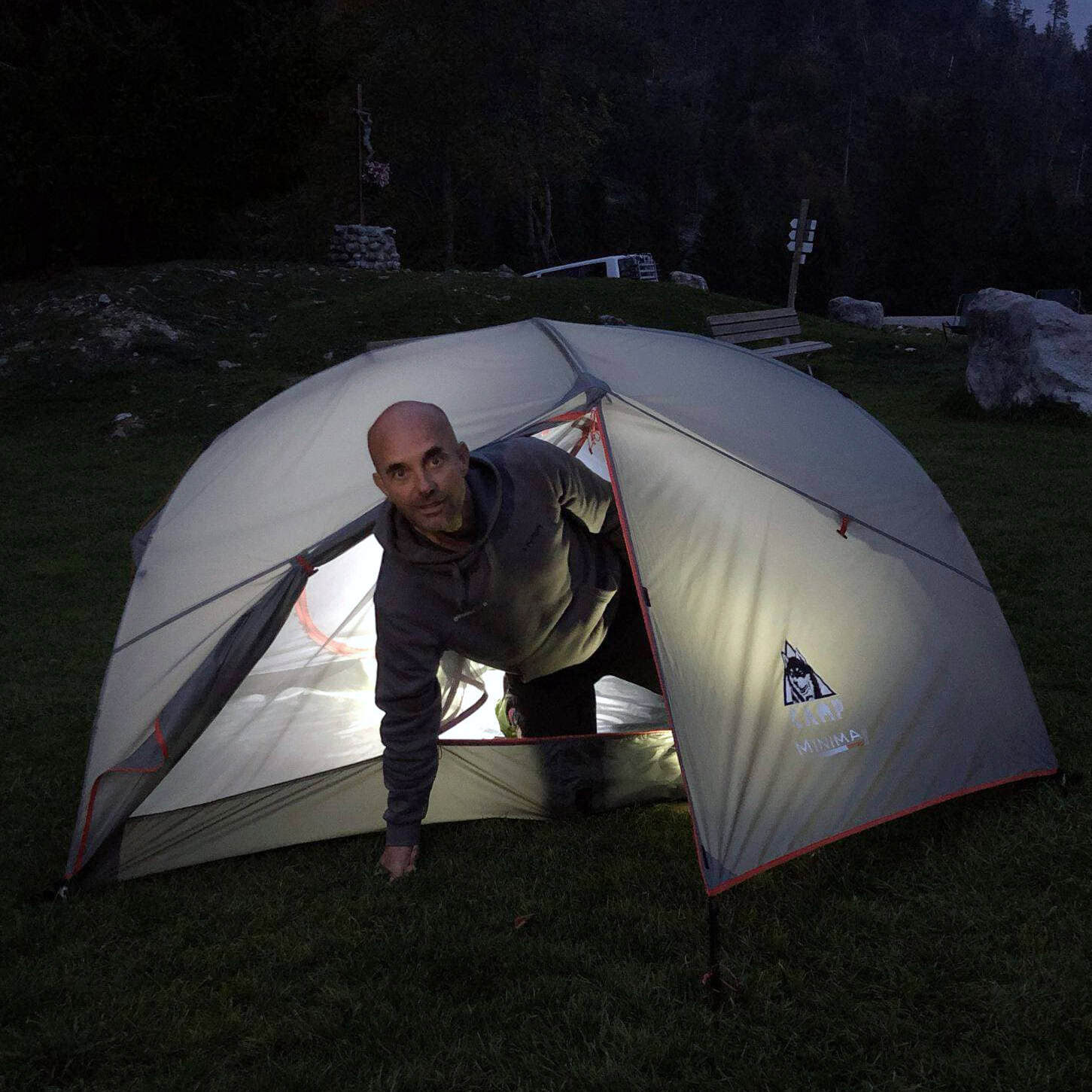 Bivacco estivo su prato alpino: tester emerge da tenda dome illuminata dall'interno al crepuscolo — attrezzatura bivacco estate 2026 Outdoortest