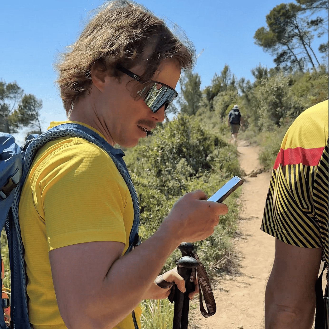 Tester Outdoortest consulta la app PeakVisor su sentiero soleggiato del Finalese, con bastoncini Leki in mano